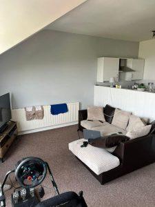 A cozy living room with a brown and beige sectional sofa, a flat-screen TV on a wooden stand, and a radiator against the wall. A steering wheel and pedal set are positioned in front of a gaming chair. This inviting space is part of Cleethorpes DN35 8LE mixed-use investment property for sale by auction.