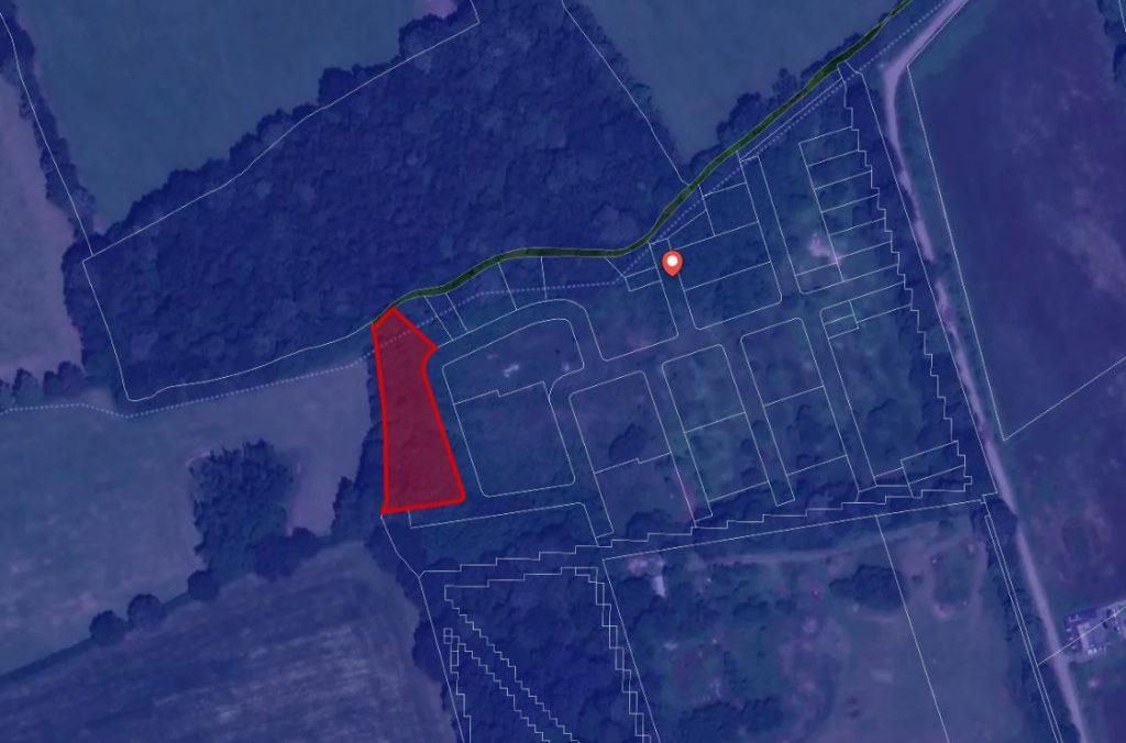 A satellite map shows a residential area with outlined plots of land. One specific plot outlined in red stands out, located near the edge of a wooded area. A main road runs near the top of the image, and a marker pin is placed near the center of the map. Land at Bones Lane sold by auction at Palace Auctions.