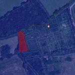 A satellite map shows a residential area with outlined plots of land. One specific plot outlined in red stands out, located near the edge of a wooded area. A main road runs near the top of the image, and a marker pin is placed near the center of the map. Land at Bones Lane sold by auction at Palace Auctions.