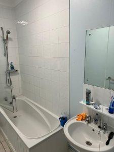 A bathroom in the mixed-use investment property for sale at 38 & 38A Alexandra Road, Cleethorpes DN35 8LE features a white bathtub and showerhead on the left, partially covered by a glass panel. The wall above the bathtub is tiled. On the right is a sink with various toiletries, including a toothbrush holder, soap dispenser, and mirror above an orange cloth.