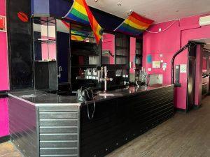 A bar with pink walls and a black counter, adorned with rainbow flags hanging from the ceiling. Located at 38 & 38A Alexandra Road in Cleethorpes DN35 8LE, this mixed-use investment property for sale by auction with Palace Auctions features mirrors, shelves behind the counter, and a quiet wooden floor.