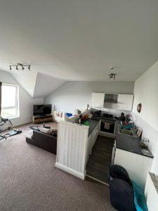 A modern, open-concept living space at 38 & 38A Alexandra Road, Cleethorpes DN35 8LE. It features a compact kitchen with white cabinets and dark countertops. Adjacent is a cozy seating area with a TV. The room has slanted ceilings, carpeting, and an exercise bike near the window. Items are on the counters and floor.