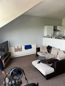 A living room at 38 & 38A Alexandra Road, Cleethorpes DN35 8LE, featuring a slanted ceiling, a brown L-shaped couch, a coffee table with a laptop, and a television in the corner. A small kitchen area and clothes drying on a radiator can be seen alongside a gaming wheel and pedals set up in front of the TV. Mixed-use investment property