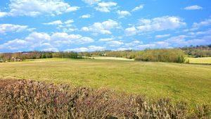 A wide-open grassy field under a bright blue sky with scattered white clouds. The landscape includes distant trees and rolling hills. A neatly trimmed hedge frames the bottom of the image. This plot of land, known as Km1 Underriver Sevenoaks, is periodically for sale with Palace Auctions and Palace Auctions London.