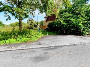 A narrow rural road bordered by greenery leads to a hidden residence partly obscured by dense trees and bushes. This unique land at Staines with future hope value for residential properties is up for sale by auction at Palace Auctions. A small pole stands near the entrance, which has a visible driveway with some uneven and cracked pavement. A wooden fence is seen in the background.