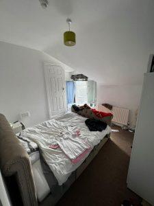A small bedroom with sloped ceilings, featuring a bed with an upholstered headboard and an assortment of clothes and items scattered on it. The room has a window with blue curtains on the far wall, a white wardrobe to the right, and a radiator beneath the window. Located at 38 & 38A Alexandra Road, Cleethorpes DN35 8LE.