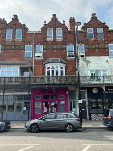 A three-story brick building at 38 & 38A Alexandra Road, Cleethorpes DN35 8LE, boasts Victorian architectural features, including arched windows and ornate trim. The ground floor houses various shops such as the bright pink storefront labeled "Silhouette." This mixed-use investment property is for sale by auction with Palace Auctions.