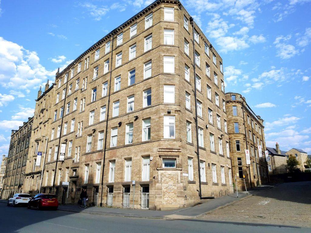 The historic Airedale House, a large six-story stone building with numerous white-framed windows, stands on a corner lot under a bright blue sky with scattered clouds. Cars are parked along the street, and a few people walk nearby. Another Bradford investment flat successfully sold at auction by Palace Auctions.