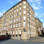 The historic Airedale House, a large six-story stone building with numerous white-framed windows, stands on a corner lot under a bright blue sky with scattered clouds. Cars are parked along the street, and a few people walk nearby. Another Bradford investment flat successfully sold at auction by Palace Auctions.