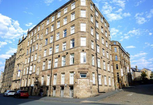 The historic Airedale House, a large six-story stone building with numerous white-framed windows, stands on a corner lot under a bright blue sky with scattered clouds. Cars are parked along the street, and a few people walk nearby. Another Bradford investment flat successfully sold at auction by Palace Auctions.