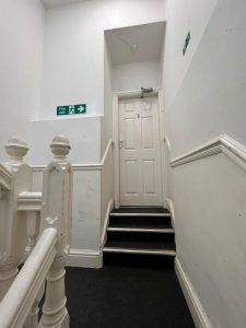 A narrow, indoor hallway with white walls and a black carpeted floor. A white, numbered door labeled "3" is at the top of a short flight of stairs. There are fire exit signs on the walls indicating the direction to the nearest exits. This mixed-use investment property at 38 & 38A Alexandra Road, Cleethorpes DN35 8LE is for sale by auction