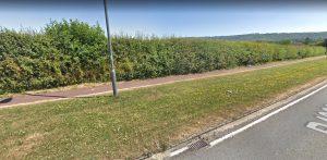 A narrow, paved pathway runs parallel to Aylesbury Road with grassy strips on both sides. Tall, dense hedges line the pathway on the left, and a metal lamppost stands along the path. Plots of land adjacent to this picturesque area are available for sale by auction with Palace Auctions. Hills with sparse vegetation are visible in the background.