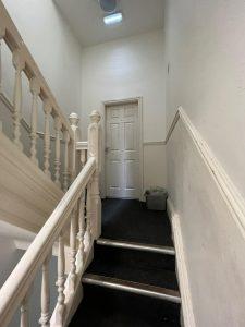 A narrow, dimly lit stairwell with white railings and grey carpet leads up to a closed, white door labeled "1" at 38 & 38A Alexandra Road, Cleethorpes DN35 8LE. A gray trash bin sits to the right of the door. The walls and ceiling are off-white, and a light fixture is on the ceiling.