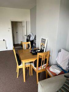 A small dining area with a wooden table and four chairs in a room. On the table are various items, including a stack of clothes and a bottle. Against the wall, there is framed artwork and a clothing item hanging on a chair. An open door leads to another room in this Cleethorpes DN35 8LE Mixed use investment property at 38 & 38A Alexandra Road for