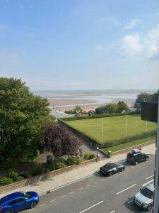 A beachfront scene taken from a higher vantage point near 38 & 38A Alexandra Road, Cleethorpes DN35 8LE. In the foreground, cars are parked along a road next to a grassy area with trees. Beyond, a sandy beach stretches toward the horizon, where the sea meets the sky under a few scattered clouds.