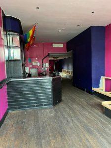 An empty room with pink and blue painted walls, a wooden floor, and a black wooden counter on the left. A rainbow flag is hanging from the ceiling near the counter. In the background, framed items lean against the walls. Located at 38 & 38A Alexandra Road, Cleethorpes DN35 8LE; mixed-use investment property for sale by auction with Palace Auctions.