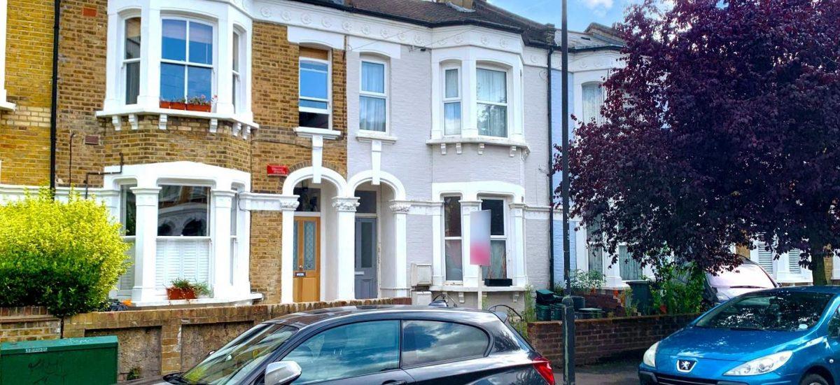 A residential street featuring traditional terraced houses with brick exteriors and white accents. Two parked cars, a black one in the foreground and a blue one on the right, are visible. A red-leaved tree partially covers the house on the right, recently sold as Flat 107 Venner Road London by Palace Auctions. The sky is partly cloudy.