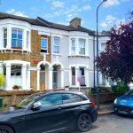 A residential street featuring traditional terraced houses with brick exteriors and white accents. Two parked cars, a black one in the foreground and a blue one on the right, are visible. A red-leaved tree partially covers the house on the right, recently sold as Flat 107 Venner Road London by Palace Auctions. The sky is partly cloudy.
