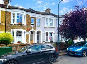 A residential street featuring traditional terraced houses with brick exteriors and white accents. Two parked cars, a black one in the foreground and a blue one on the right, are visible. A red-leaved tree partially covers the house on the right, recently sold as Flat 107 Venner Road London by Palace Auctions. The sky is partly cloudy.