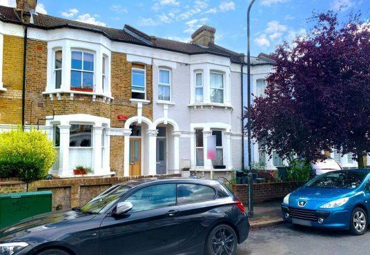 A residential street featuring traditional terraced houses with brick exteriors and white accents. Two parked cars, a black one in the foreground and a blue one on the right, are visible. A red-leaved tree partially covers the house on the right, recently sold as Flat 107 Venner Road London by Palace Auctions. The sky is partly cloudy.