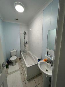 A compact bathroom with light blue walls at 38 & 38A Alexandra Road, Cleethorpes DN35 8LE. It features a white bathtub with a showerhead, a toilet, and a pedestal sink. Above the sink is a rectangular mirror. The floor is tiled, and bath mats are placed in front of the bathtub and toilet. Various toiletries are visible.