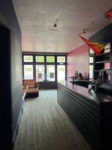 Image of a dimly lit bar with a long counter on the right, a person sitting at the end of the counter, and booths on the left. Rainbow flags hang from the ceiling, and colorful stained-glass windows provide natural light in the background. Located at 38 & 38A Alexandra Road, Cleethorpes DN35 8LE. Mixed-use investment property for sale by auction