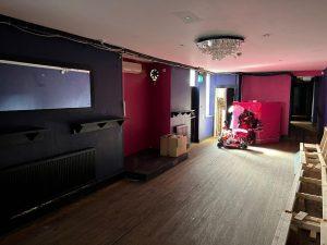 A dimly lit room with dark purple and pink walls sits within the mixed-use investment property at 38 & 38A Alexandra Road, Cleethorpes DN35 8LE. The wooden floor is scattered with a few cardboard boxes, while a mirror and shelves adorn the left wall. In the rear, a red and black children's ride-on car is illuminated under a chandelier, awaiting its future