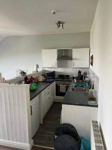 A compact kitchen at 38 & 38A Alexandra Road, Cleethorpes DN35 8LE, features white cabinets and dark countertops. The space includes a stove with a range hood, a sink, and various kitchen items like containers and a drying rack. White tiles adorn the wall next to a small lamp by the stove. Mixed-use investment property for sale by auction with Palace Auctions