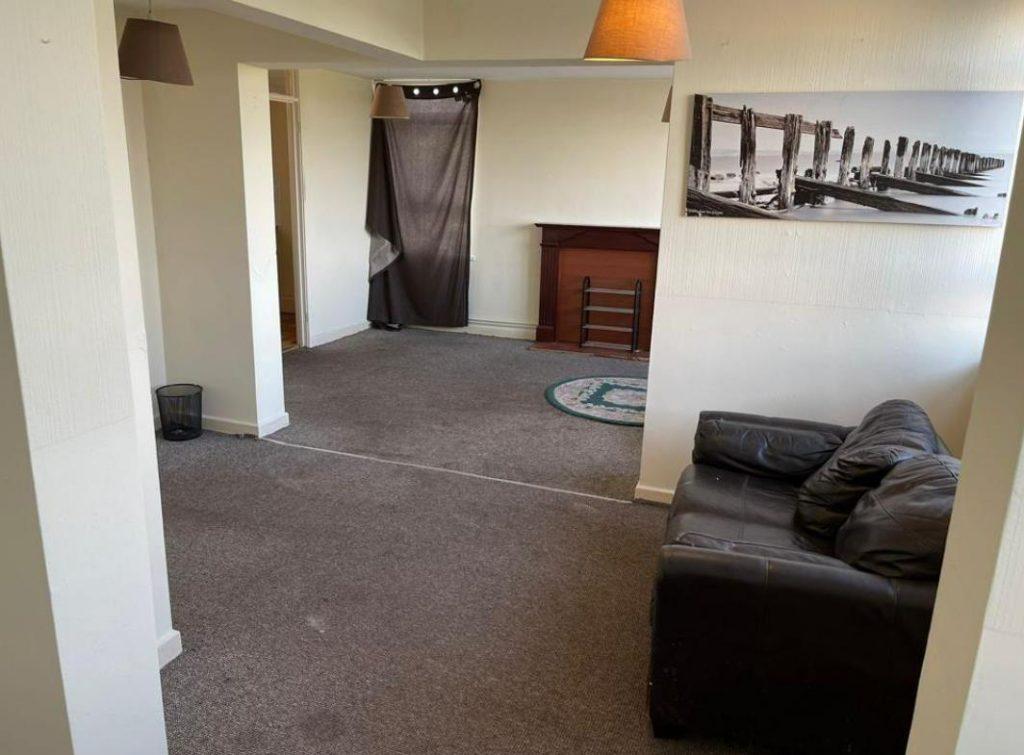 A living room in Collingwood Court with beige walls and carpet. A dark brown leather sofa is positioned near a white wall, and there is a wooden fireplace with a dark screen. A black and white photo hangs on the wall above. Brown curtains cover a window, and a round rug is on the floor. Investment apartments for sale by auction with Palace Auctions.