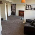 A living room in Collingwood Court with beige walls and carpet. A dark brown leather sofa is positioned near a white wall, and there is a wooden fireplace with a dark screen. A black and white photo hangs on the wall above. Brown curtains cover a window, and a round rug is on the floor. Investment apartments for sale by auction with Palace Auctions.