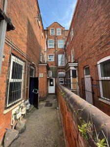 A narrow alleyway between 38 & 38A Alexandra Road, Cleethorpes DN35 8LE, leads to a rear entrance. The brick buildings have several windows, some covered with bars. The sky is clear and blue. A tall wall with green plants growing at the top lines the right side of the alley. Mixed-use investment property for sale by auction with Palace Auctions.
