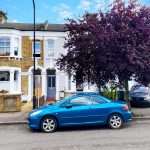 A blue car is parked on a residential street in front of a row of two-story brick houses with white accents. Nearby, a large tree with purple foliage stands to the right, partially obscuring a black car. The sky is partly cloudy over this serene neighborhood where Flat 107 Venner Road London was successfully sold by Palace Auctions.