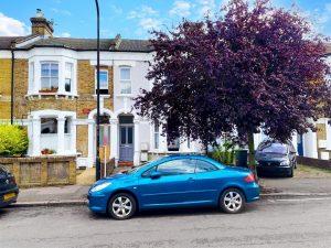 A blue car is parked on a residential street in front of a row of two-story brick houses with white accents. Nearby, a large tree with purple foliage stands to the right, partially obscuring a black car. The sky is partly cloudy over this serene neighborhood where Flat 107 Venner Road London was successfully sold by Palace Auctions.