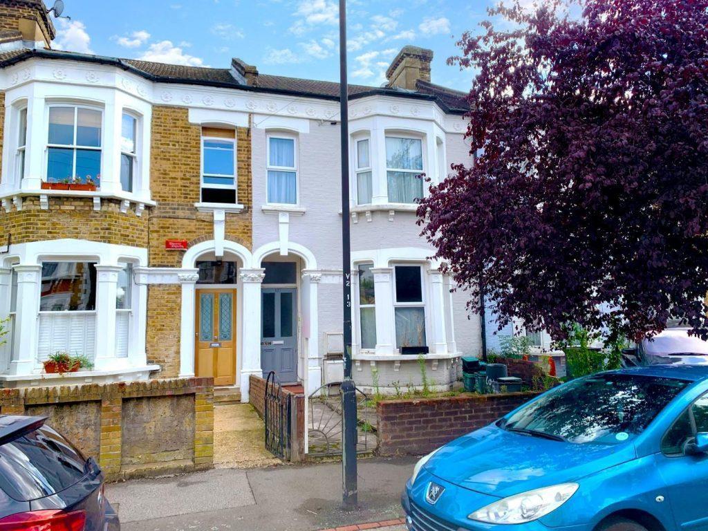 Two adjacent Victorian-style terraced houses with bay windows and ornate detailing stand poised on Flat 107 Venner Road, London. The left house is yellow brick with a wooden front door, the right is light gray with a gray door. A blue car is parked in front, beside a reddish-purple tree. Successfully sold by Palace Auctions.