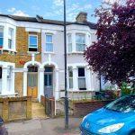 Two adjacent Victorian-style terraced houses with bay windows and ornate detailing stand poised on Flat 107 Venner Road, London. The left house is yellow brick with a wooden front door, the right is light gray with a gray door. A blue car is parked in front, beside a reddish-purple tree. Successfully sold by Palace Auctions.