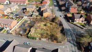 Property for sale by Auction An aerial view of Cradley Heath captures a residential neighborhood with pitched-roof houses, paved roads, and parked cars. The image centers on development land at Peartree—a fenced, overgrown lot with dense vegetation—contrasting sharply with the well-maintained properties surrounding it. Presented by Palace Auctions
