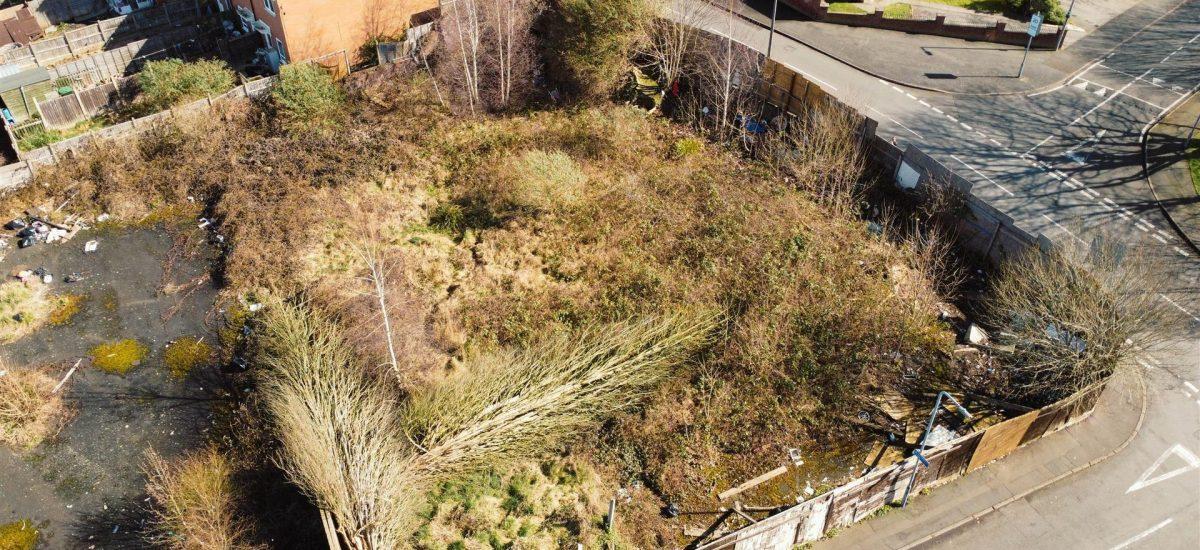 Property for sale by Auction Aerial view of an overgrown vacant lot surrounded by a wooden fence in Cradley Heath. The lot, potential development land at Peartree, has patches of tall grasses, bushes, and some garbage. Nearby are brick houses and intersecting streets with sidewalks. The area appears neglected and unkempt. Presented by Palace Auctions