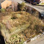Property for sale by Auction Aerial view of an overgrown vacant lot surrounded by a wooden fence in Cradley Heath. The lot, potential development land at Peartree, has patches of tall grasses, bushes, and some garbage. Nearby are brick houses and intersecting streets with sidewalks. The area appears neglected and unkempt. Presented by Palace Auctions
