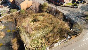 Property for sale by Auction Aerial view of an overgrown vacant lot surrounded by a wooden fence in Cradley Heath. The lot, potential development land at Peartree, has patches of tall grasses, bushes, and some garbage. Nearby are brick houses and intersecting streets with sidewalks. The area appears neglected and unkempt. Presented by Palace Auctions