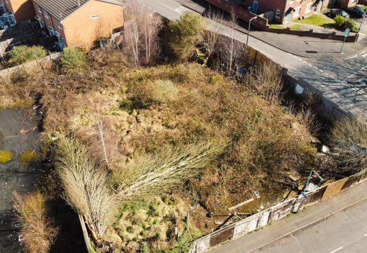 Property for sale by Auction Aerial view of an overgrown vacant lot surrounded by a wooden fence in Cradley Heath. The lot, potential development land at Peartree, has patches of tall grasses, bushes, and some garbage. Nearby are brick houses and intersecting streets with sidewalks. The area appears neglected and unkempt. Presented by Palace Auctions