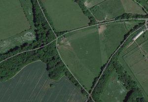 Property for sale by Auction Aerial view of a rural area with fields, trees, and a few roads. Pebble Lane and Crampshaw Lane intersect on the right side, near the outskirts of Ashtead. The landscape is predominantly green with scattered patches of woodland and agricultural fields. Buildings are sparse. Presented by Palace Auctions