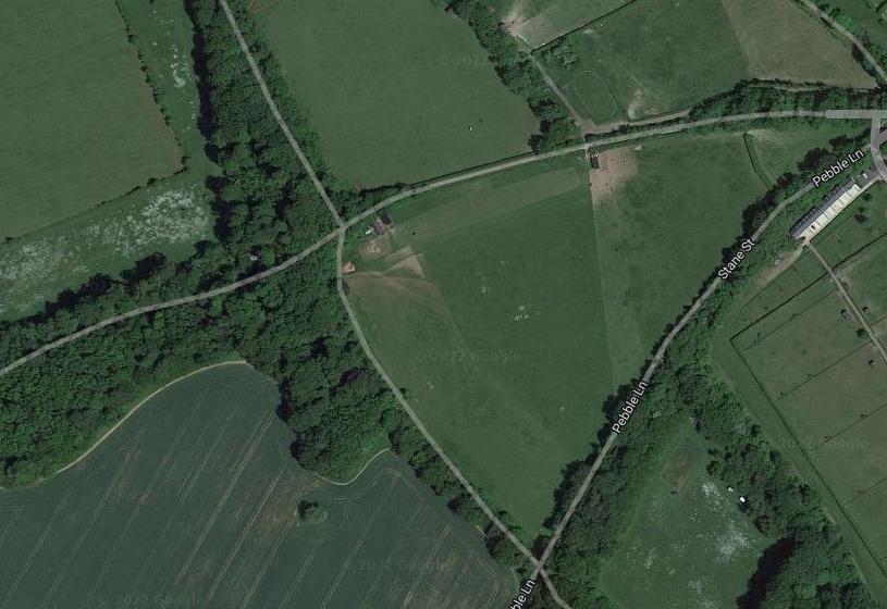 Property for sale by Auction Aerial view of a rural area with fields, trees, and a few roads. Pebble Lane and Crampshaw Lane intersect on the right side, near the outskirts of Ashtead. The landscape is predominantly green with scattered patches of woodland and agricultural fields. Buildings are sparse. Presented by Palace Auctions
