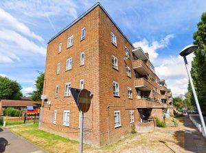 Property for sale by Auction A three-story brick apartment building on a sunny day at 21 Glazebrook Close, Dulwich. The building features several balconies with railings and numerous windows. A sidewalk runs along the building, and a street lamp is visible. A street sign and trees frame the quintessential London scene. Presented by Palace Auctions