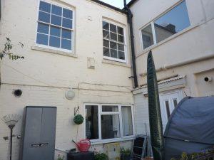 Property for sale by Auction A white-painted, two-story house at 3-4 Western Road with large rectangular windows. The ground floor features a grey shed, a red watering can, a green topiary, and garden furniture. A rake is leaning against the wall. A gray tent or covered object is seen on the right near the Edingburugh mews TN37 6DG. Presented by Palace Auctions