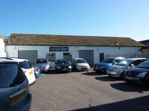 Property for sale by Auction A group of cars is parked in front of a single-story, white building with a sign that reads "WE HAVE MOVED" to 3-4 Western Road & the Edingburugh mews TN37 6DG. The building has a worn roof and two large closed garage doors against the backdrop of a clear blue sky. Presented by Palace Auctions