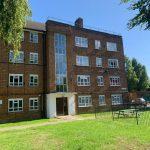 Property for sale by Auction A brick apartment building at 21 Glazebrook Close in Dulwich features multiple floors with numerous windows and a central section adorned with vertical glass panes. In front, a grassy lawn encloses a fenced area containing a picnic table and bench. Trees and a clear blue sky complete the serene backdrop. Presented by Palace Auctions