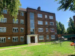 Property for sale by Auction A brick apartment building at 21 Glazebrook Close in Dulwich features multiple floors with numerous windows and a central section adorned with vertical glass panes. In front, a grassy lawn encloses a fenced area containing a picnic table and bench. Trees and a clear blue sky complete the serene backdrop. Presented by Palace Auctions