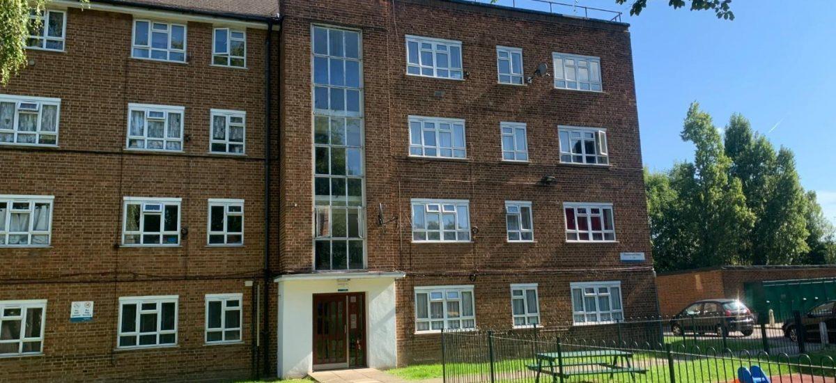 Property for sale by Auction A brick apartment building at 21 Glazebrook Close, Dulwich, London, with four stories and numerous windows is set against a clear blue sky. In front, there's a small playground area with a slide, grass lawn, and a few benches. Trees and a fence are also visible around the perimeter of the building. Presented by Palace Auctions