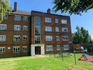 Property for sale by Auction A brick apartment building at 21 Glazebrook Close, Dulwich, London, with four stories and numerous windows is set against a clear blue sky. In front, there's a small playground area with a slide, grass lawn, and a few benches. Trees and a fence are also visible around the perimeter of the building. Presented by Palace Auctions