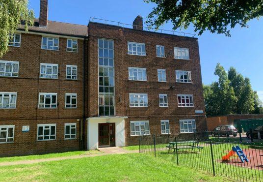Property for sale by Auction A brick apartment building at 21 Glazebrook Close, Dulwich, London, with four stories and numerous windows is set against a clear blue sky. In front, there's a small playground area with a slide, grass lawn, and a few benches. Trees and a fence are also visible around the perimeter of the building. Presented by Palace Auctions
